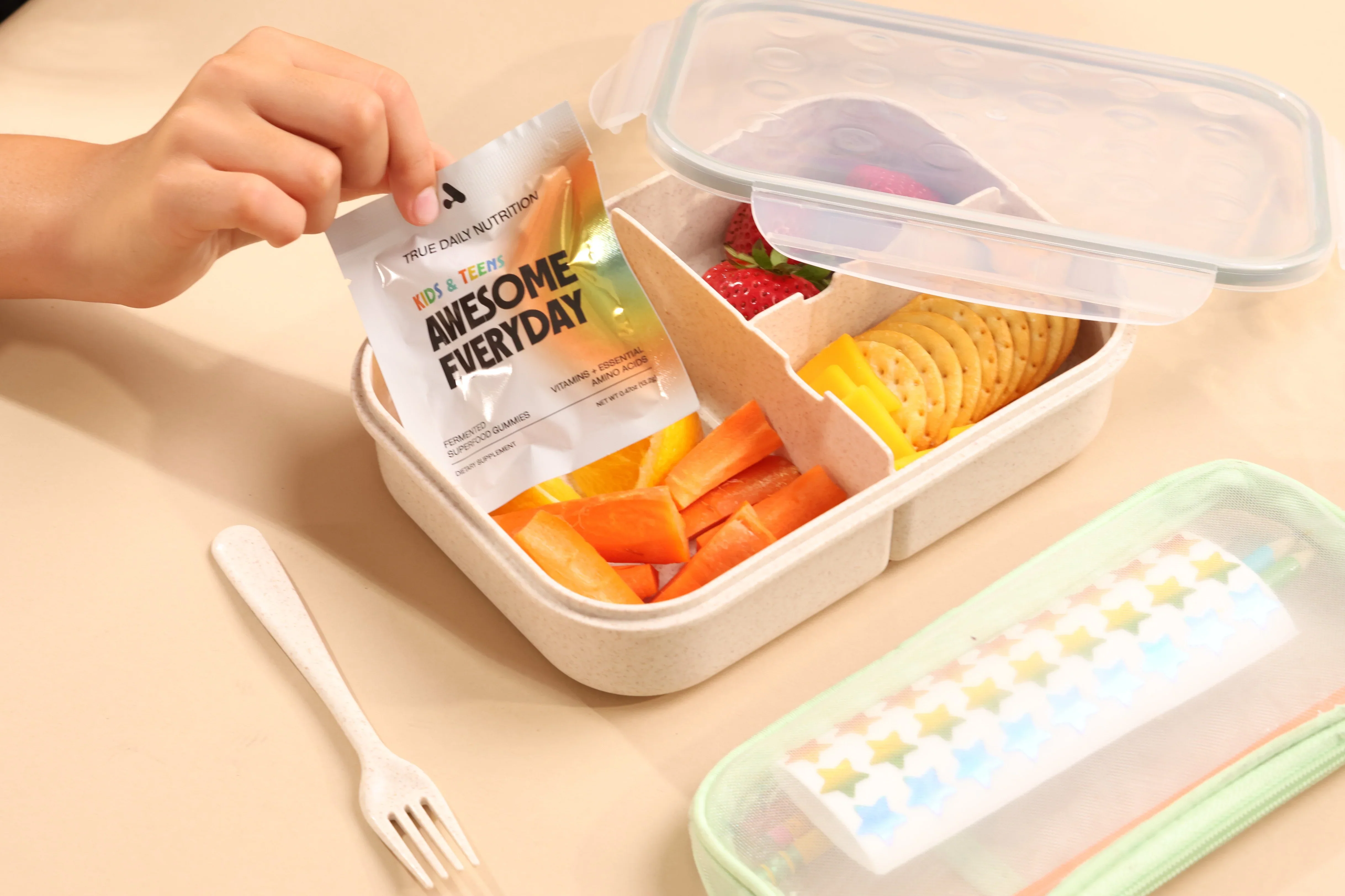 Awesome Everyday™ Kids & Teens packet placed beside a child's lunchbox with fruit, crackers, and vegetables.