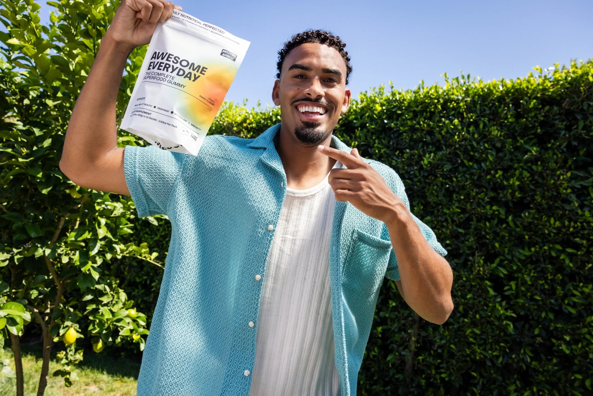 Man holding an Awesome Everyday gummy packet outdoors beside green hedges.